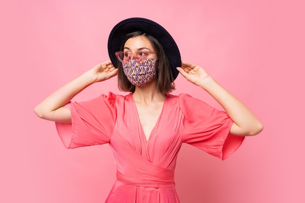 Fashionable woman dressed protective stylish face mask. Wearing black hat and sunglasses. Posing over pink wall