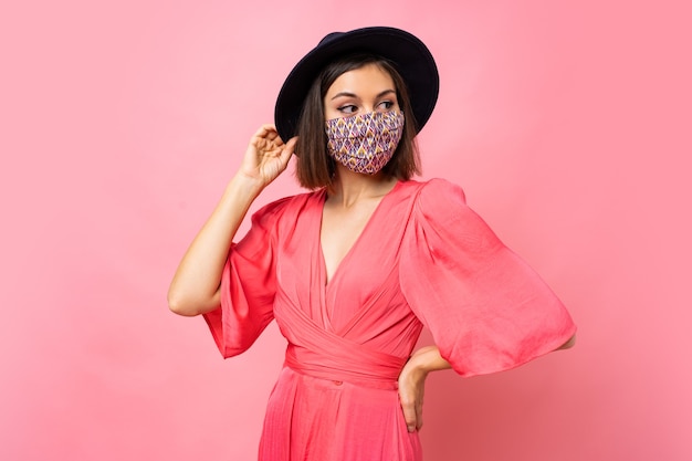 Fashionable woman dressed protective stylish face mask. Wearing black hat and sunglasses. Posing over pink wall