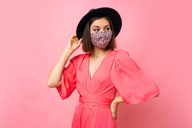 Fashionable woman dressed protective stylish face mask. Wearing black hat and sunglasses. Posing over pink wall