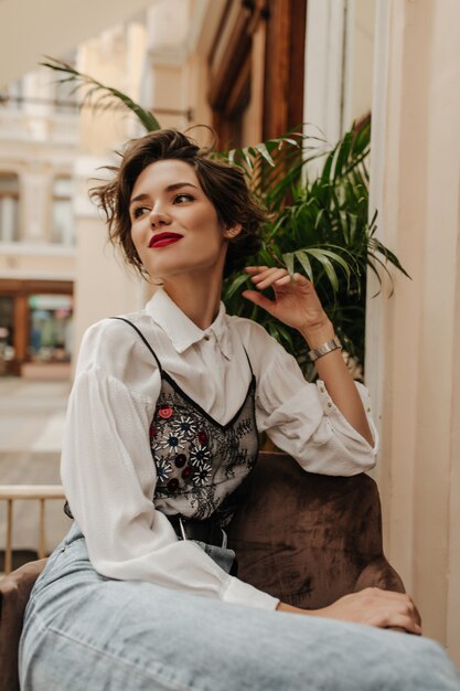 Fashionable woman in white blouse and jeans smiling in cafe. Trendy woman with short brunette hair sitting in restaurant.