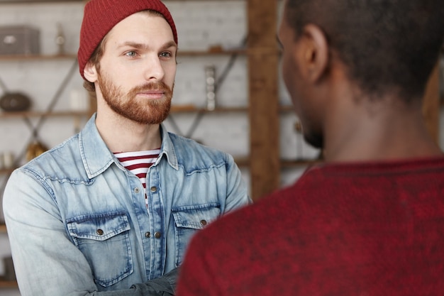 Fashionable young bearded Caucasian man wearing trendy hat and denim shirt smiling while having nice conversation with unrecognizable dark-skinned man during meeting in modern restaurant interior