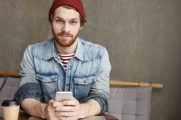 Fashionable young bearded male in hat and denim shirt sitting at cafe table with paper cup of fresh coffee, holding mobile phone while messaging online and surfing internet, using free wi-fi