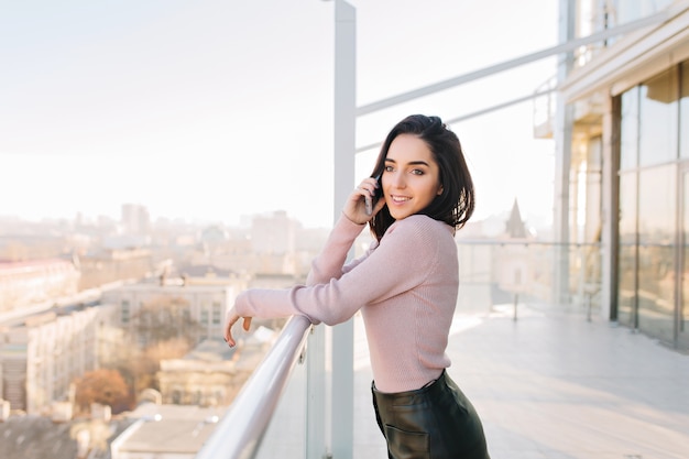 Free photo fashionable young brunette woman speaking on phone on terrace on city view in sunny morning. success, luxury lifestyle, chilling, relaxing, joyful businesswoman, smiling to side.