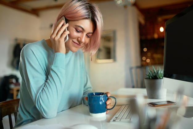 Free photo fashionable young female copywriter with pink hair and facial piercing sitting at her workplace in front of desktop computer, holding cup, drinking coffee and having phone conversation, smiling