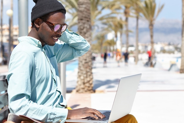 Fashionable young freelancer wearing stylish clothes and accessories sitting on bench