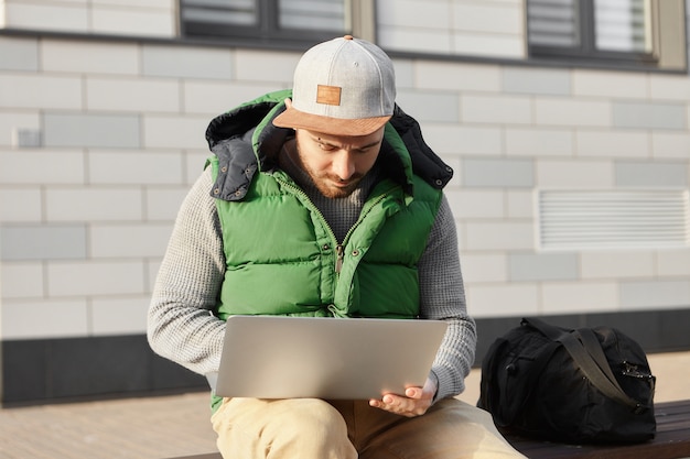 Free photo fashionable young man traveler sitting on bench with bag, holding portable computer on his lap, booking apartment while staying in foreign city on business trip. technology, traveling and lifestyle