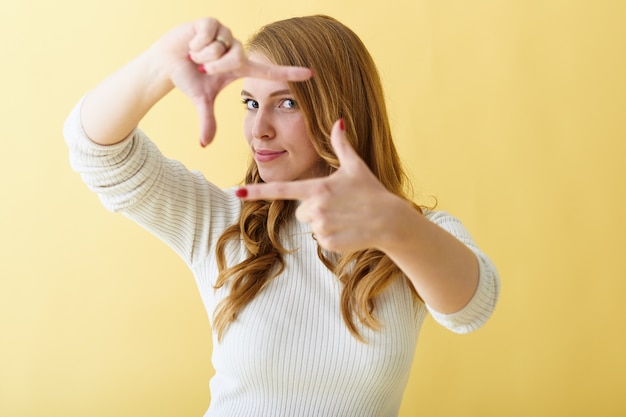 Free photo fashionable young positive lady with red manicured nails gesturing, making camera frame with her fingers, posing isolated against blank yellow copy space wall background for your text or advertisement