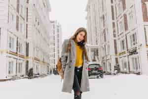 Free photo fashionable young woman in coat with backpack walking on street in big city in snowing time. cheerful mood, snowfall, waiting for christmas, expressing positivity, true emotions.