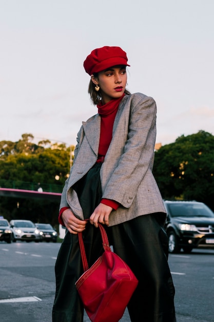 Free photo fashionable young woman in red cap posing on road holding handbag