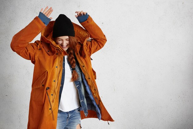 Free photo fashionable young woman wearing red winter coat, black hat and ragged jeans raising hands in air