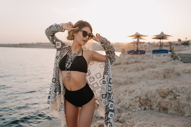 Free photo fasionable model posing on beach, sunset. sexy girl wearing black bikini, swimsuit with hight waist, cardigan, cape with ornaments , beautiful beach, sea, rock.