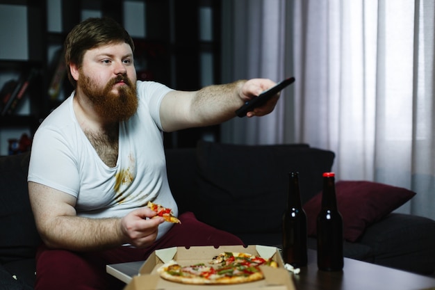 Fat man eats pizza sitting on the sofa and switches channels on the TV-set