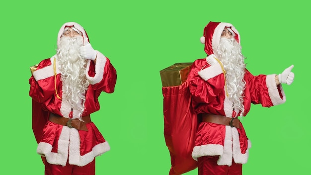 Free photo father christmas shows thumbs up on camera feeling positive giving like and agreement symbol over greenscreen backdrop santa claus in suit expressing approval with gifts bag