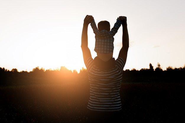 Free Photo father and son walking on the field at the sunset time, boy sitting on mans shoulders. 