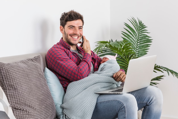 Free photo father with baby talking on phone and using laptop