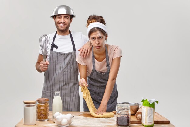 Fatigue woman kneads raw dough, makes bread, busy with baking pastry, cheerful man stands near, dressed in apron, holds whisk, tries to help. Two cooks bake confectionery at home, try new recipe.