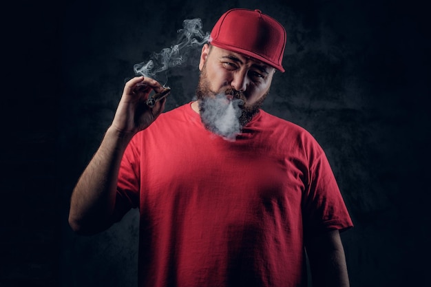 Fatty bearded hipster male in a red hip hop clothes smoking a cigarette on a grey background.