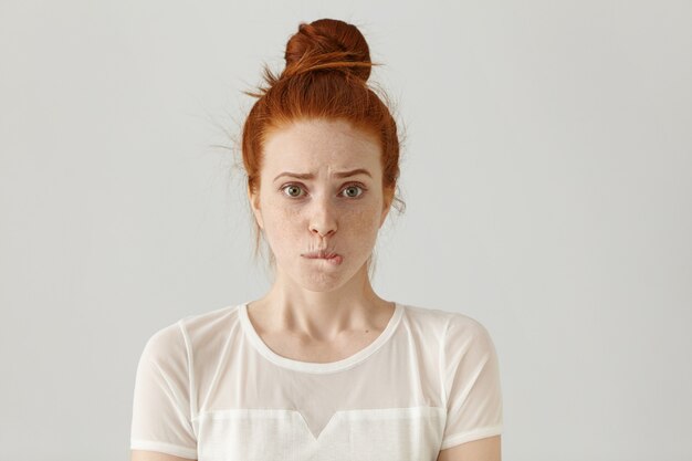 Fearful young Caucasian female with ginger hair dressed in white blouse having confused guilty look, biting her lower lip, feeling sorry for doing something wrong and making terrible mistake