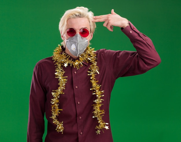 Free Photo fed up young blonde man wearing glasses and protective mask with tinsel garland around neck looking at camera doing suicide gesture isolated on green background