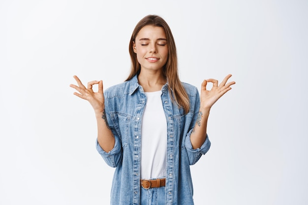 Free photo feeling zen in body. relaxed young woman standing calm and peaceful in meditating pose, close eyes and breathing, standing on white