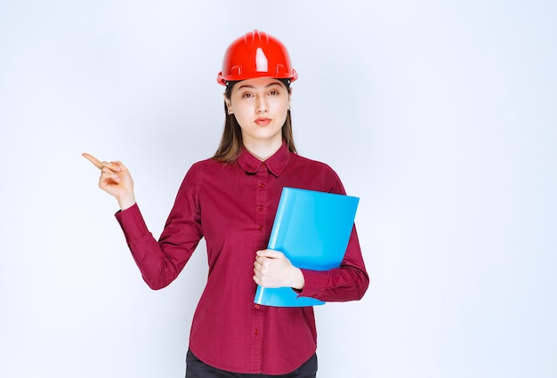 Female architect in red helmet with blue folder getting standing and posing. 