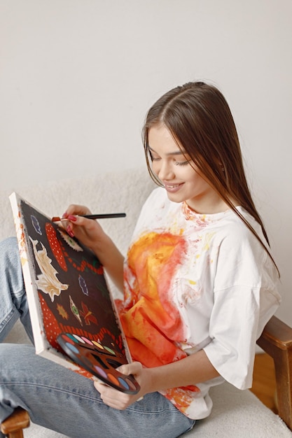 Female artist sitting on a chair near white wall holding a canvas and brushes