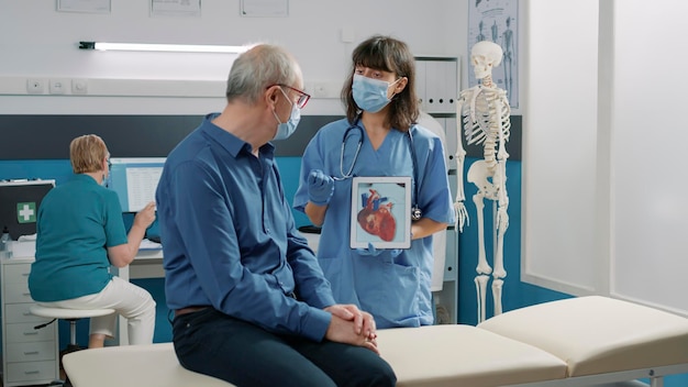 Free photo female assistant holding tablet with cardiovascular system illustration, explaining heart condition to retired person during covid 19 pandemic. nurse showing cardiology diagnosis on gadget.