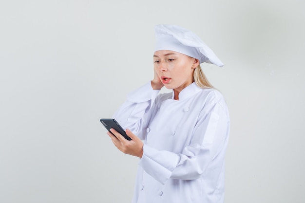 Female chef holding smartphone with hand on head in white uniform and looking surprised.