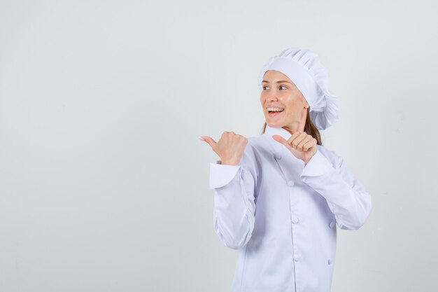 Female chef pointing thumbs to side in white uniform and looking cheerful