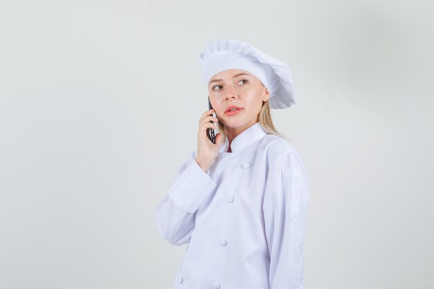 Female chef talking on smartphone and looking aside in white uniform .