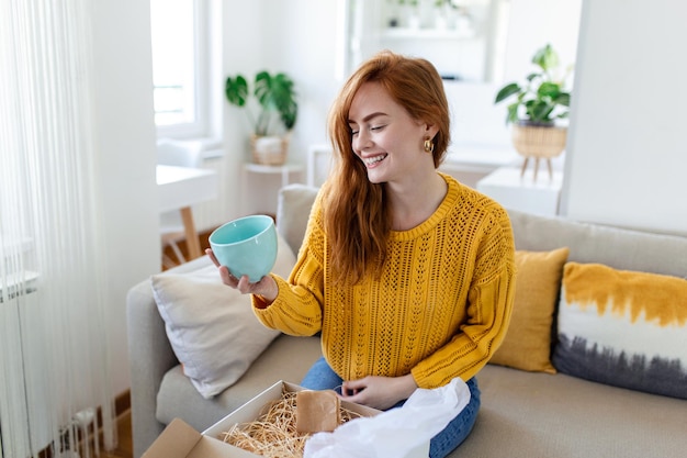 Female consumer unpack parcel receive retail purchase fast postal shipping delivery concept Beautiful young woman is holding cardboard box sitting on sofa at home