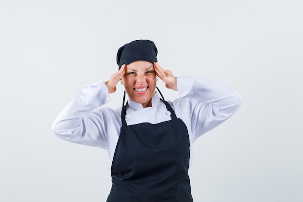 Female cook holding hands on temples in uniform, apron and looking cheery. front view.