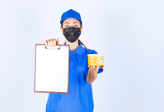 Female courier in blue uniform and face mask delivering a takeaway food and asking the customer to sign. 