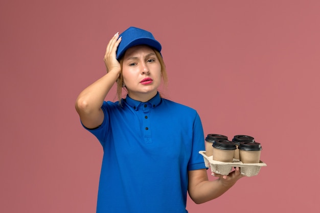 female courier in blue uniform holding brown delivery cups of coffee having headache on the pink, service worker uniform delivery