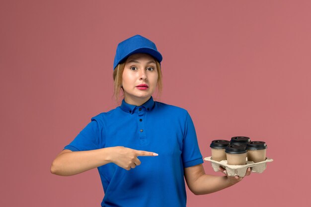 female courier in blue uniform holding brown delivery cups of coffee pointing out on pink, service uniform delivery job worker