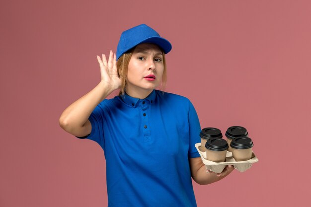 female courier in blue uniform holding brown delivery cups of coffee trying to hear out on pink, service uniform delivery worker