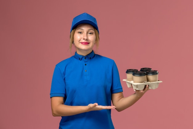 female courier in blue uniform holding brown delivery cups of coffee with slight smile on pink, service uniform delivery job