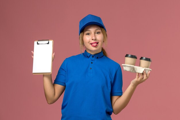 female courier in blue uniform holding delivery cups of coffee and notepad with tongue on pink, service worker uniform delivery