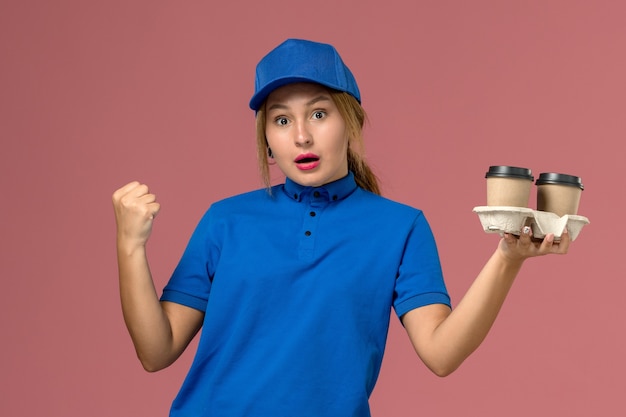 female courier in blue uniform holding delivery cups of coffee posing on pink, service worker job uniform delivery