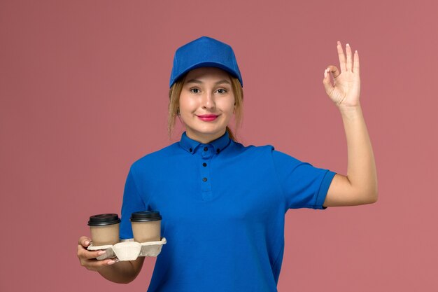 female courier in blue uniform holding delivery cups of coffee smiling on pink, service uniform delivery