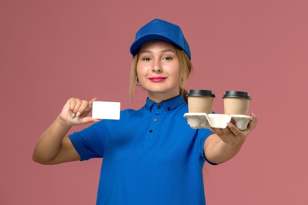 female courier in blue uniform holding delivery cups of coffee and white card on pink, service uniform delivery worker