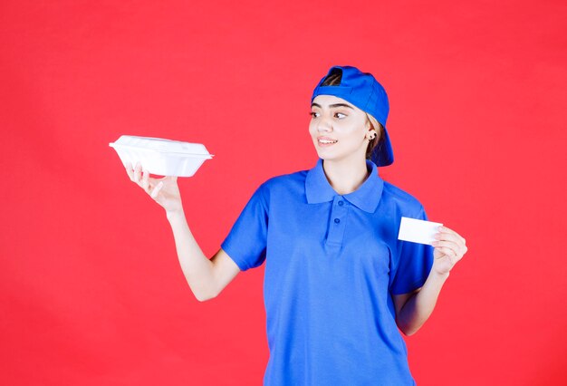 Female courier in blue uniform holding a white takeaway box and presenting her business card. 