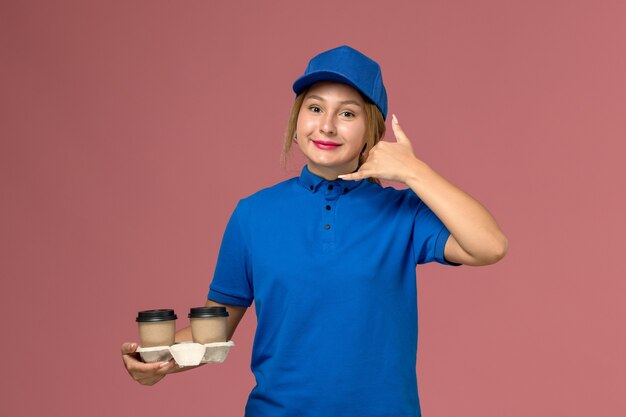 female courier in blue uniform posing and holding cups of coffee showing phone call gesture on pink, service uniform delivery job