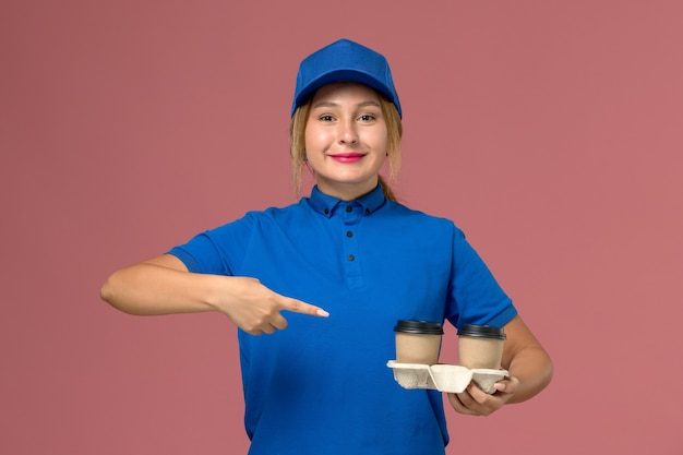 female courier in blue uniform posing holding cups of coffee with slight smile on pink, service uniform delivery job worker