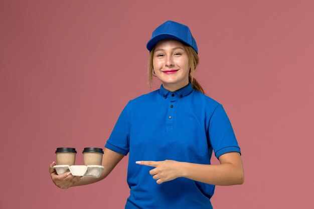 female courier in blue uniform posing and holding cups of coffee with a smile on pink, service uniform delivery job