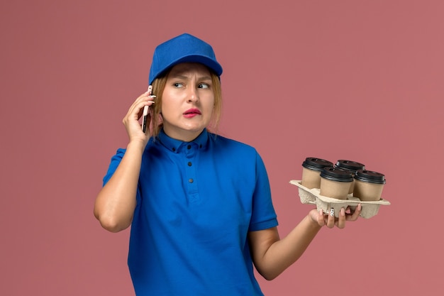 female courier in blue uniform talking on phone and holding brown cups of coffee on light-pink, work service uniform delivery