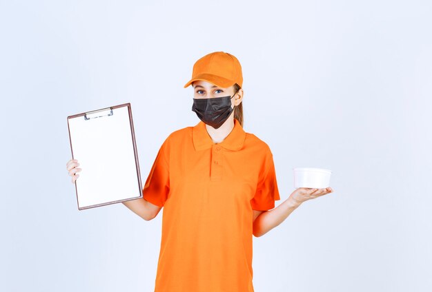 Free photo female courier in orange uniform and black mask holding a plastic cup and customer list