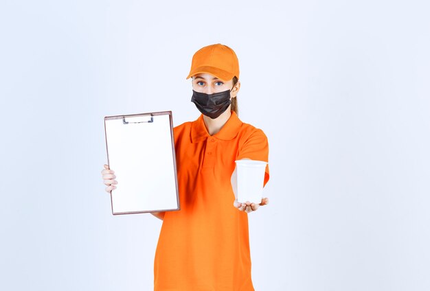 Female courier in orange uniform and black mask holding a plastic takeaway cup and presenting the signature blank for handover