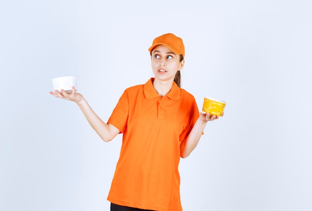 Female courier in orange uniform holding plastic and yellow noodles cup in both hands.