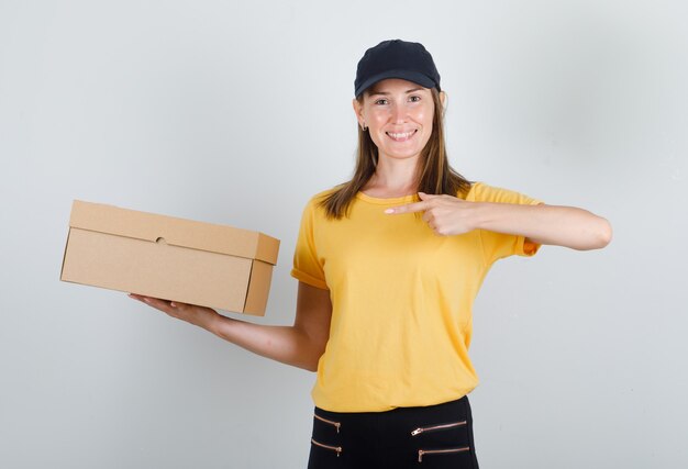 Female courier pointing finger at cardboard box in t-shirt, pants, cap and looking glad